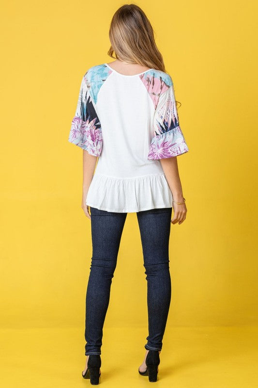 A woman stands against a yellow background wearing the Mix Pattern Circular Flounce Sleeve Tunic in navy, showcasing floral and polka dot designs on the sleeves.