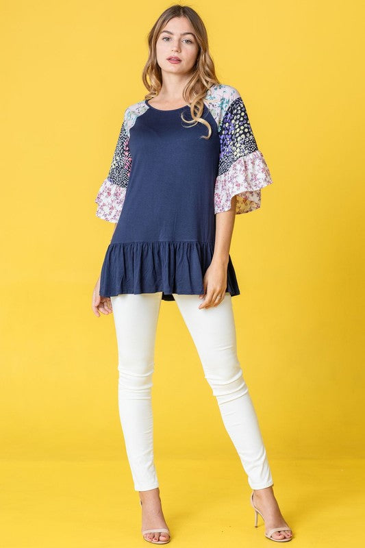 A woman stands against a yellow background wearing the Mix Pattern Circular Flounce Sleeve Tunic in navy, showcasing floral and polka dot designs on the sleeves.