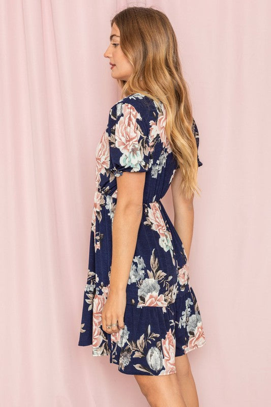 Dressed in a Surplice Tiered Midi Dress, a woman elegantly showcases the navy fabric decorated with pink and white floral patterns set against a delicate light pink background. Crafted from a cozy polyester spandex blend, this dress, made in the United States, highlights exceptional quality and sophistication.