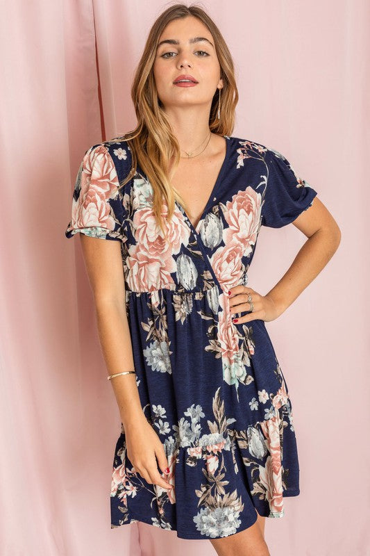 Dressed in a Surplice Tiered Midi Dress, a woman elegantly showcases the navy fabric decorated with pink and white floral patterns set against a delicate light pink background. Crafted from a cozy polyester spandex blend, this dress, made in the United States, highlights exceptional quality and sophistication.