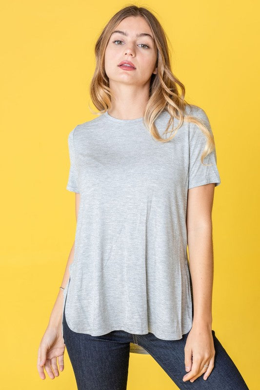 Dressed in a vibrant purple t-shirt, a woman stands against a pink backdrop. Her white pants perfectly complement the Solid Side Slit High Low Tunic style she's wearing. She accessorizes with a long necklace and gives a slight smile.