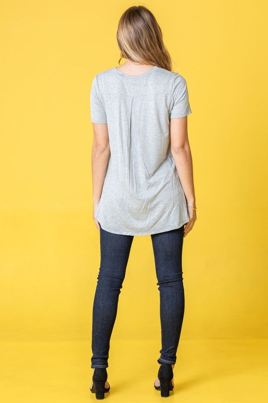 A person wearing a Solid Side Slit High Low Tunic in light blue and jeans poses gracefully against a pink curtain. Made in the United States, this look seamlessly combines casual comfort with stylish elegance.