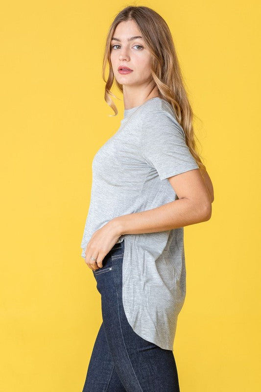 Dressed in a vibrant purple t-shirt, a woman stands against a pink backdrop. Her white pants perfectly complement the Solid Side Slit High Low Tunic style she's wearing. She accessorizes with a long necklace and gives a slight smile.