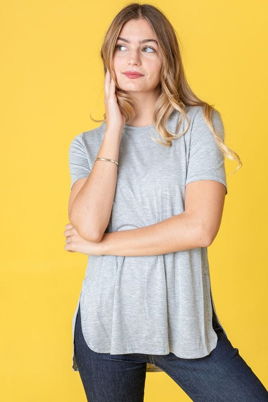 Dressed in a vibrant purple t-shirt, a woman stands against a pink backdrop. Her white pants perfectly complement the Solid Side Slit High Low Tunic style she's wearing. She accessorizes with a long necklace and gives a slight smile.