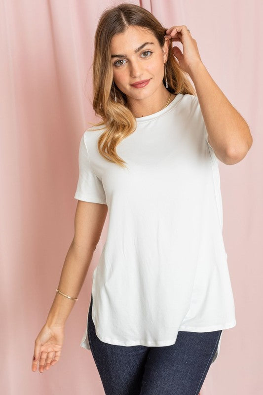 Dressed in a vibrant purple t-shirt, a woman stands against a pink backdrop. Her white pants perfectly complement the Solid Side Slit High Low Tunic style she's wearing. She accessorizes with a long necklace and gives a slight smile.