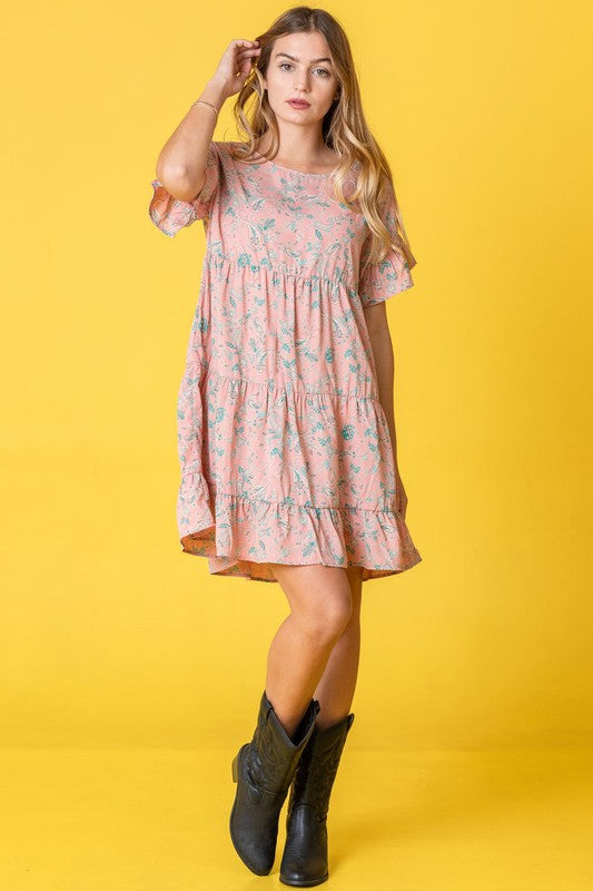 A woman in a Tiered Ruffle Trim Shift Dress stands against a pink backdrop, playfully holding a strand of her hair.