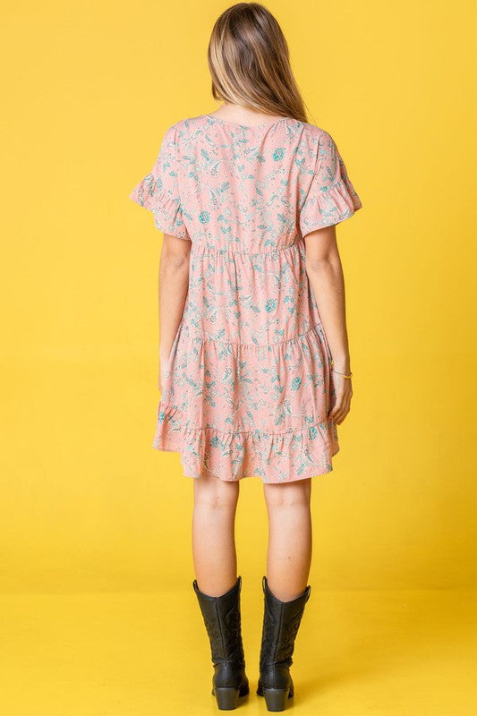 A woman in a Tiered Ruffle Trim Shift Dress featuring pink florals and a delicate paisley pattern, set against a yellow background, brushing her hair back with one hand.