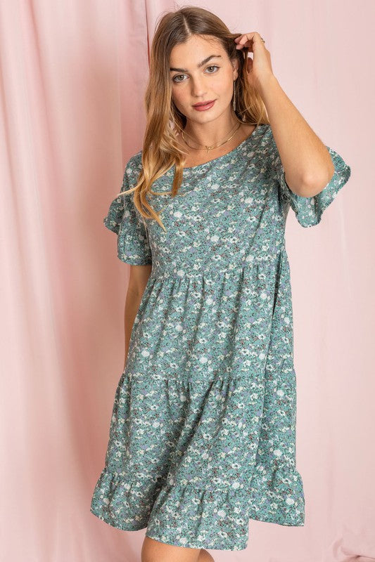 A woman in a Tiered Ruffle Trim Shift Dress featuring pink florals and a delicate paisley pattern, set against a yellow background, brushing her hair back with one hand.