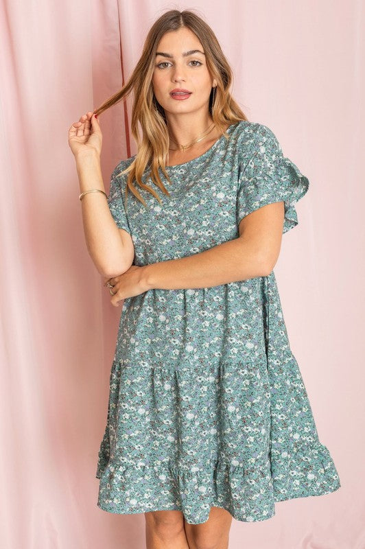 A woman in a Tiered Ruffle Trim Shift Dress featuring pink florals and a delicate paisley pattern, set against a yellow background, brushing her hair back with one hand.