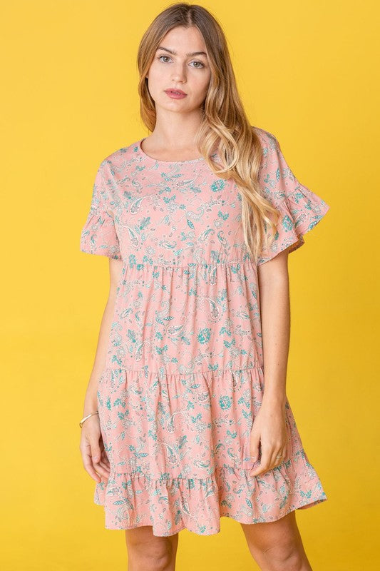 A woman in a Tiered Ruffle Trim Shift Dress featuring pink florals and a delicate paisley pattern, set against a yellow background, brushing her hair back with one hand.