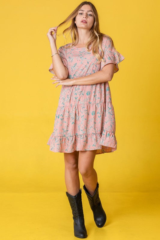 A woman in a Tiered Ruffle Trim Shift Dress featuring pink florals and a delicate paisley pattern, set against a yellow background, brushing her hair back with one hand.