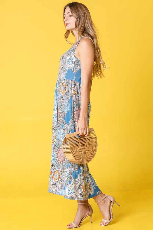 A person wearing a Sleeveless Print Ruffled Hem Tea-Length Dress stands against a pink curtain backdrop, their look reminiscent of a delicate medallion in bloom.