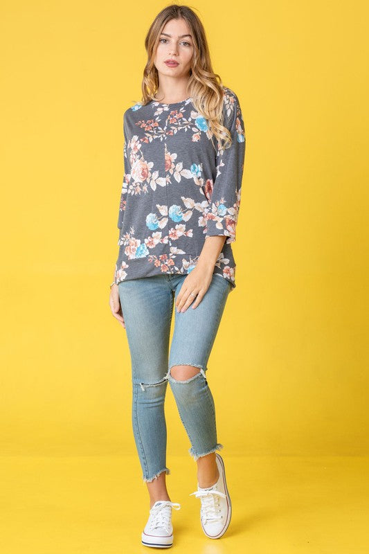 A woman with long hair wearing the Plus Spring Floral Tunic and blue jeans stands against a light pink background. She is holding her hair with one hand, capturing the essence of spring.