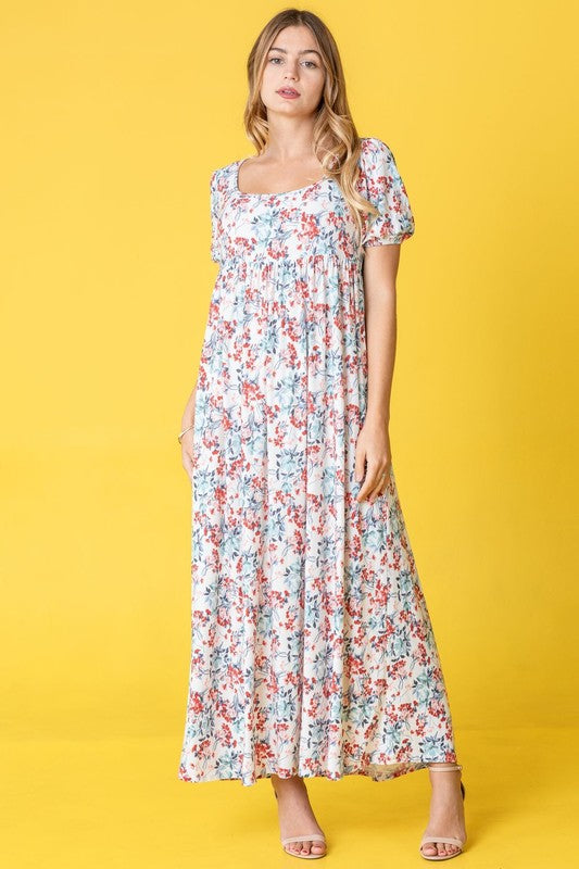 A person stands gracefully in front of a pink backdrop, wearing the Floral Square Neck Puff Sleeve dress, a blue maxi adorned with charming floral patterns.