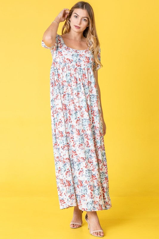 A person stands gracefully in front of a pink backdrop, wearing the Floral Square Neck Puff Sleeve dress, a blue maxi adorned with charming floral patterns.