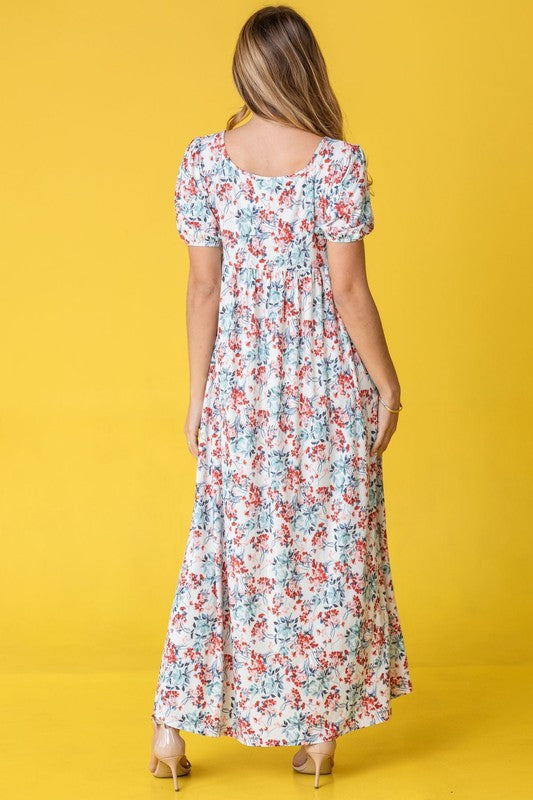 A person stands gracefully in front of a pink backdrop, wearing the Floral Square Neck Puff Sleeve dress, a blue maxi adorned with charming floral patterns.