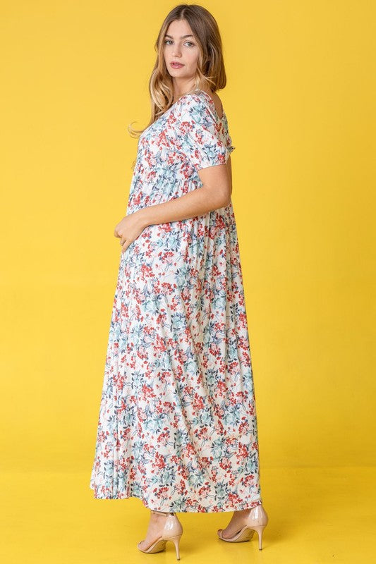 A person stands gracefully in front of a pink backdrop, wearing the Floral Square Neck Puff Sleeve dress, a blue maxi adorned with charming floral patterns.