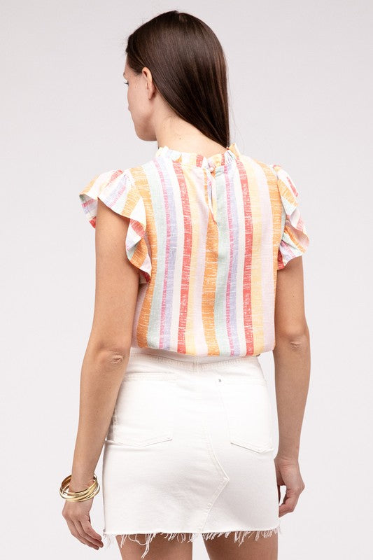 A woman sits on a white chair, wearing the Multi Stripe Ruffle Top made of non-stretch fabric and white shorts, with her hands resting on her knee. Crafted from polyester for easy care, this top maintains its vibrant colors even after a machine wash cold.