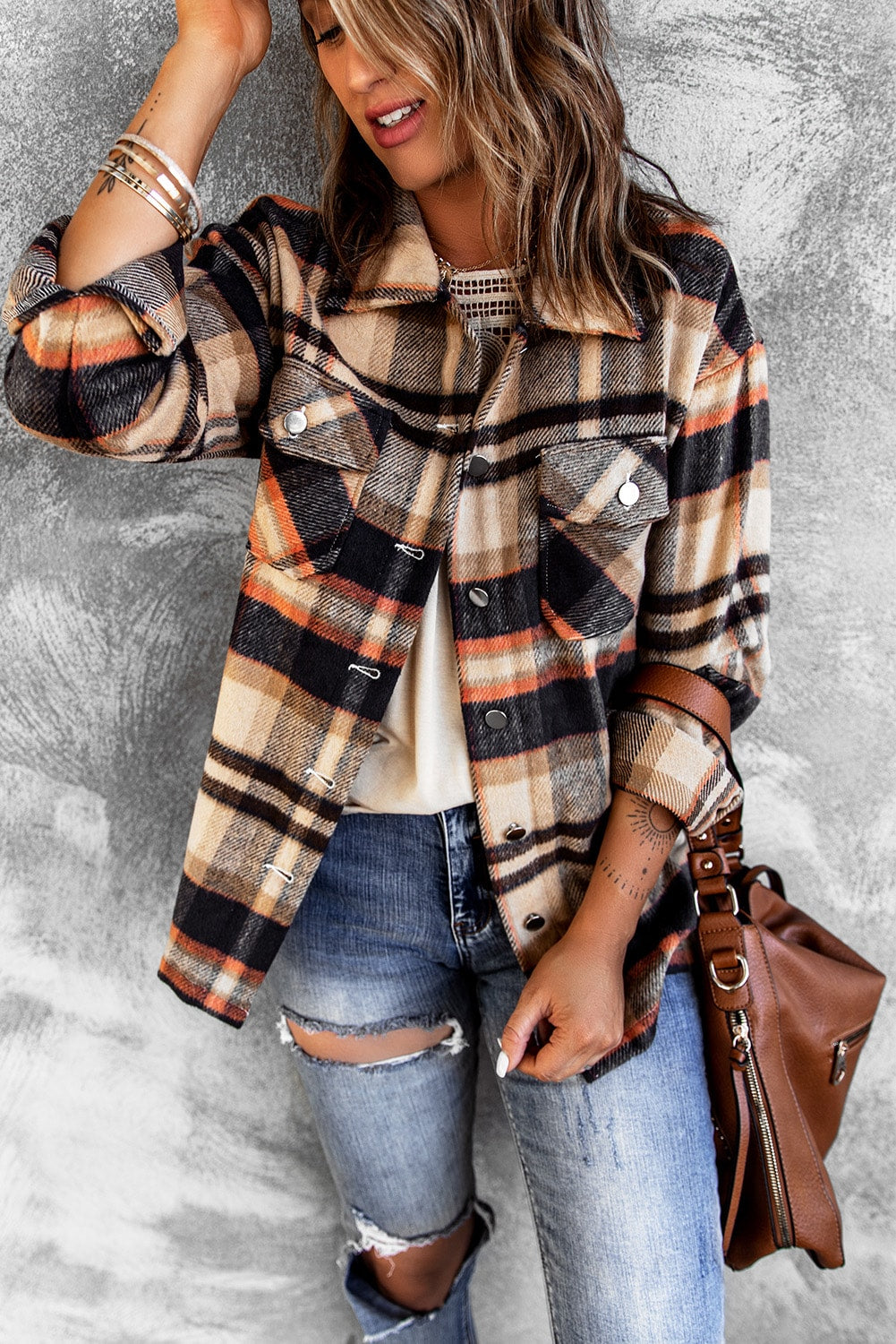 A person wearing a Brown Plaid Button Up Long Sleeve Flannel Shacket layered over a white top with ripped jeans and holding a brown handbag stands against a textured gray wall.