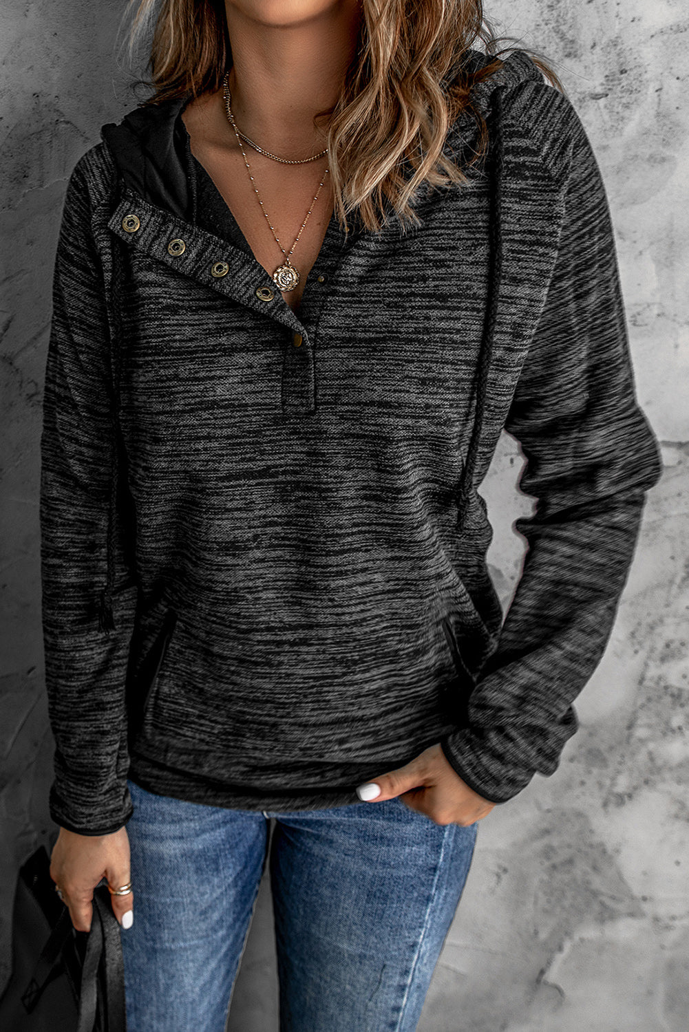 Person with wavy hair, wearing a stylish Black Heathered Print Button Snap Neck Pullover Hoodie and blue jeans, standing with their back facing the camera against a gray textured wall.