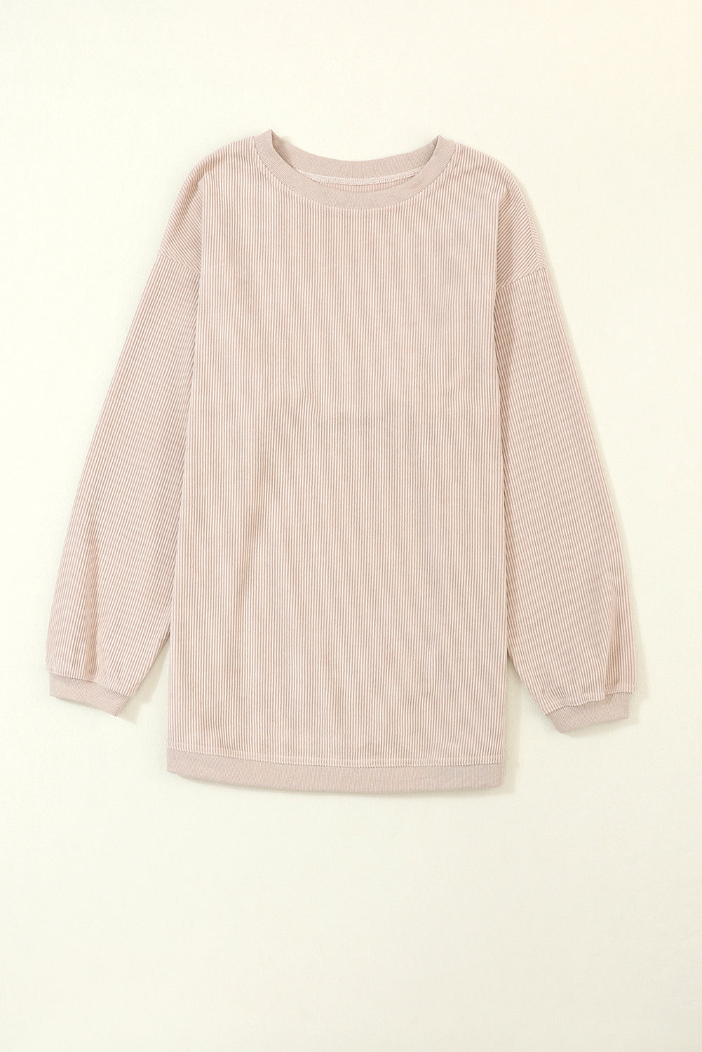 A woman stands outdoors, looking down while touching her hair with one hand. She is wearing an Apricot Ribbed Corduroy Oversized Sweatshirt and white pants, exuding an effortlessly chic and fashionable look.