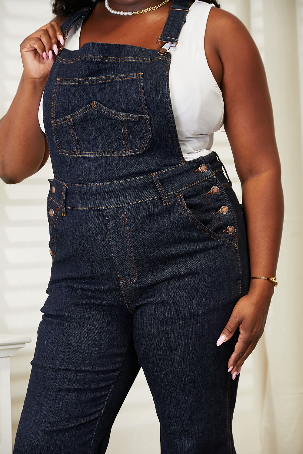 A person stands indoors wearing Judy Blue Full Size High Waist Classic Denim Overalls and a white tank top, with one hand in the pocket and the other resting on a white chest of drawers, next to a vase of dried pampas grass.