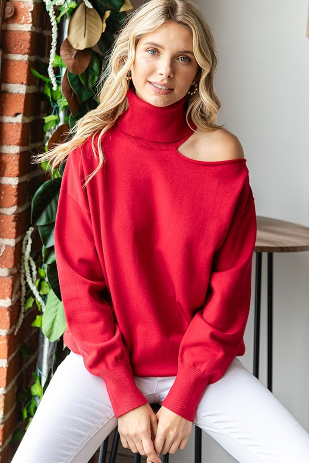 A blonde woman in the stylish First Love Cut Out Shoulder Turtleneck Sweater and white pants sits on a stool against a wall, with plants in the background.