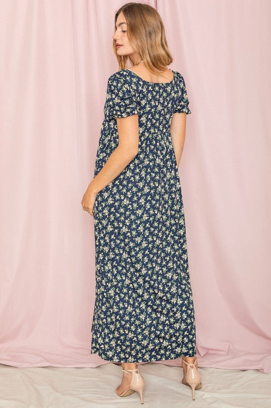 A person stands gracefully in front of a pink backdrop, wearing the Floral Square Neck Puff Sleeve dress, a blue maxi adorned with charming floral patterns.