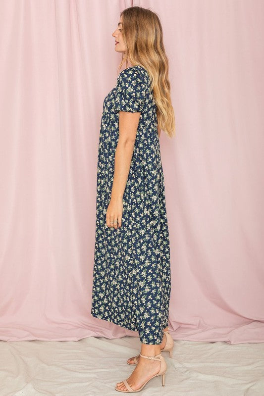 A person stands gracefully in front of a pink backdrop, wearing the Floral Square Neck Puff Sleeve dress, a blue maxi adorned with charming floral patterns.