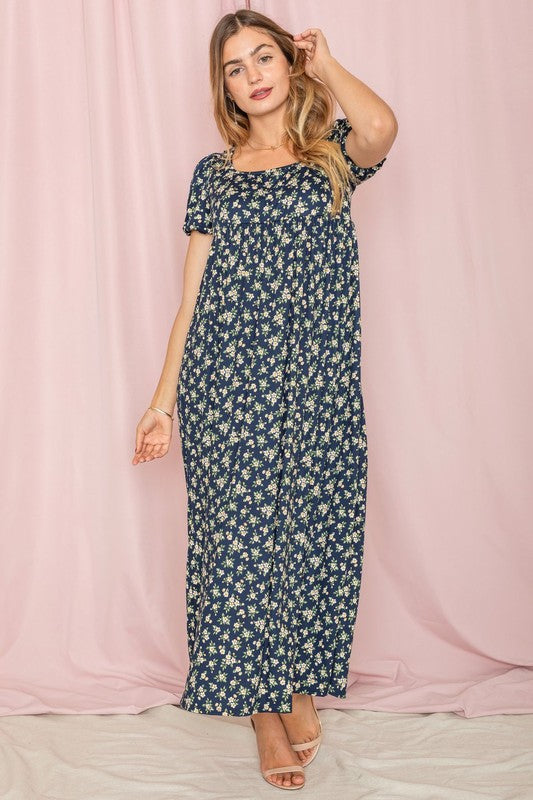 A person stands gracefully in front of a pink backdrop, wearing the Floral Square Neck Puff Sleeve dress, a blue maxi adorned with charming floral patterns.