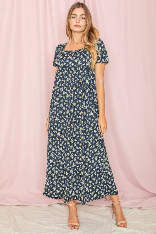 A person stands gracefully in front of a pink backdrop, wearing the Floral Square Neck Puff Sleeve dress, a blue maxi adorned with charming floral patterns.