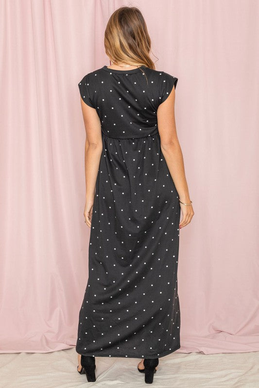 A woman in a Polka Dot Folded Cap Sleeve Maxi Dress and white sneakers stands gracefully before a pink curtain. Made in the United States, this stylish outfit blends elegance with casual flair.