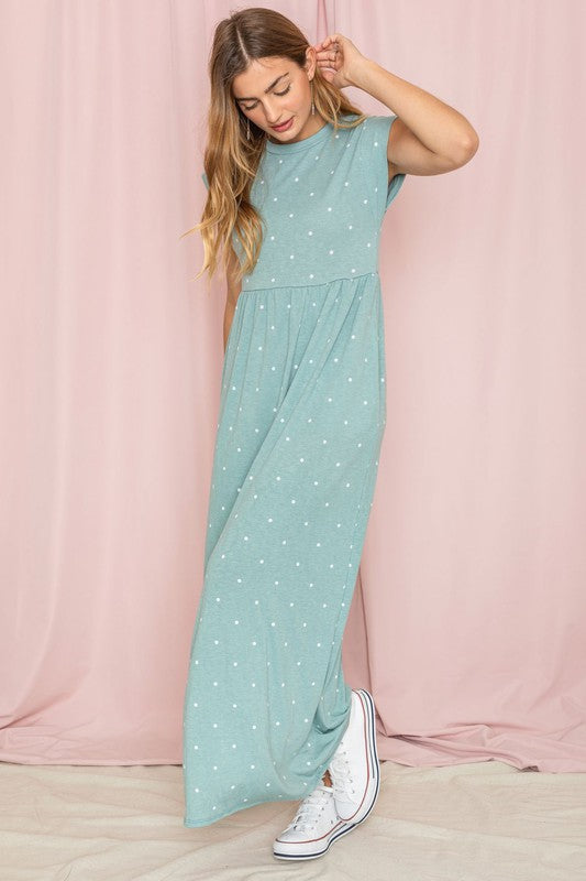 A woman in a Polka Dot Folded Cap Sleeve Maxi Dress and white sneakers stands gracefully before a pink curtain. Made in the United States, this stylish outfit blends elegance with casual flair.