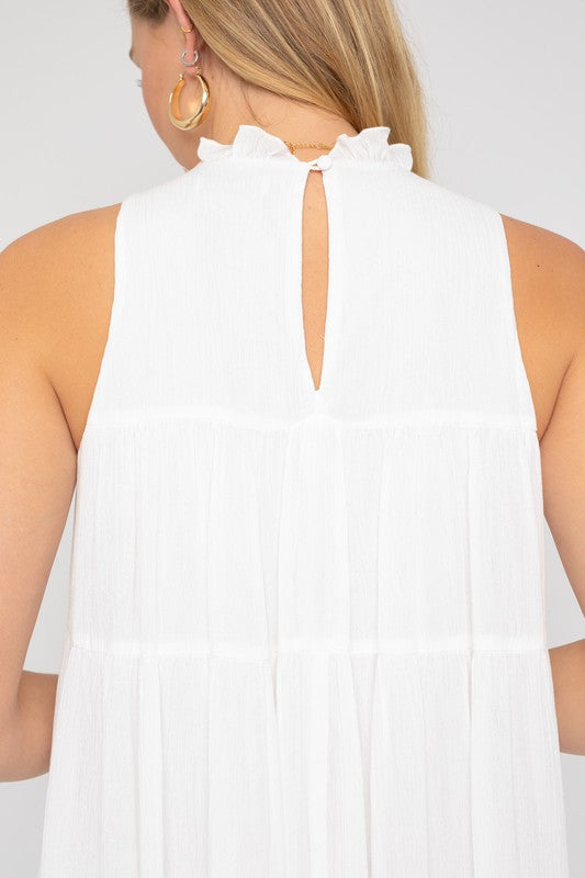 A woman smiles and looks down while wearing the Sleeveless Ruffle Neck Tiered Dress, holding a small brown purse against a plain background.