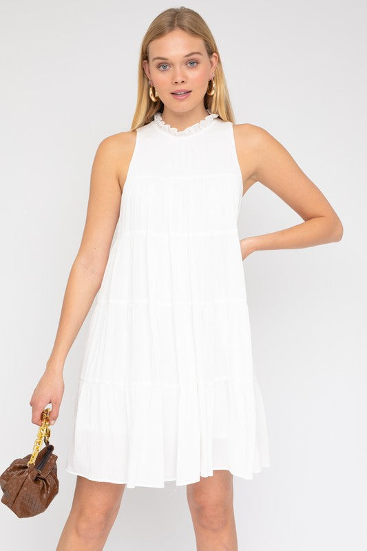 A woman smiles and looks down while wearing the Sleeveless Ruffle Neck Tiered Dress, holding a small brown purse against a plain background.