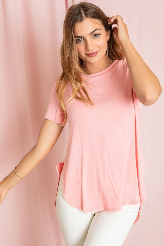 A person wearing a Solid Side Slit High Low Tunic in light blue and jeans poses gracefully against a pink curtain. Made in the United States, this look seamlessly combines casual comfort with stylish elegance.