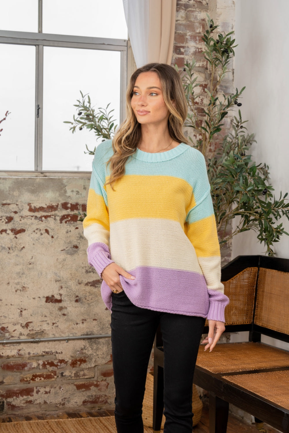 A person stands indoors near a window, wearing the Sew In Love Full Size Color Block Exposed Seam Sweater and black pants. The cozy knitwear complements the rustic walls and nearby plant, adding a touch of warmth to the scene.