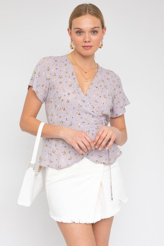 A woman in a light purple S/S Wrap W/Self Waist Tie Ditsy Print Top and white skirt, holding a white handbag, stands against a plain background.