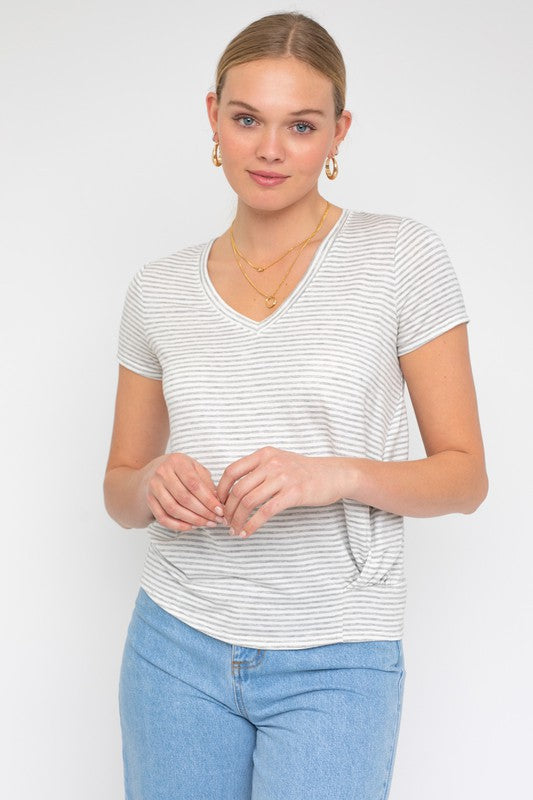 A person wearing a Short Sleeve V-Neck Twist Front Top in gray and white stripes, paired with blue jeans, poses with a smile against a plain background.