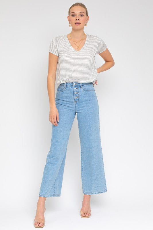 A person wearing a Short Sleeve V-Neck Twist Front Top in gray and white stripes, paired with blue jeans, poses with a smile against a plain background.