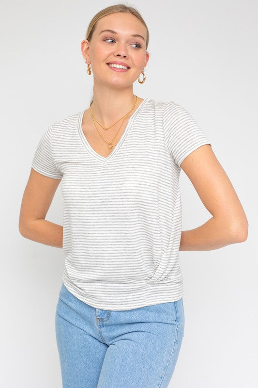 A person wearing a Short Sleeve V-Neck Twist Front Top in gray and white stripes, paired with blue jeans, poses with a smile against a plain background.