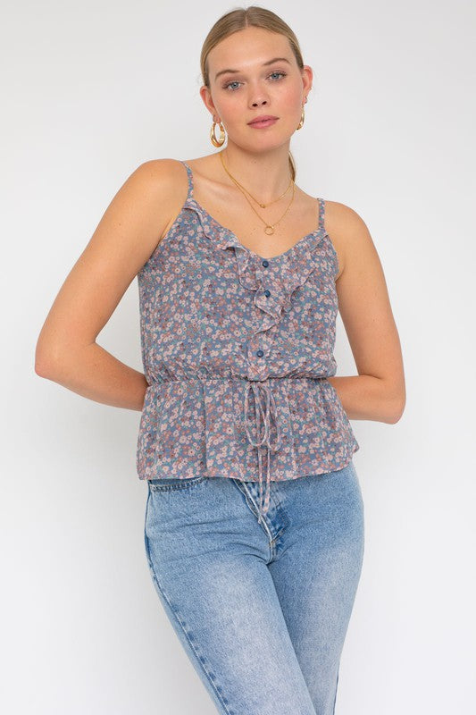 A person wearing a Sleeveless Elastic Waist Top With Ruffle, showcasing a floral pattern, paired with light blue jeans, stands against a white background.