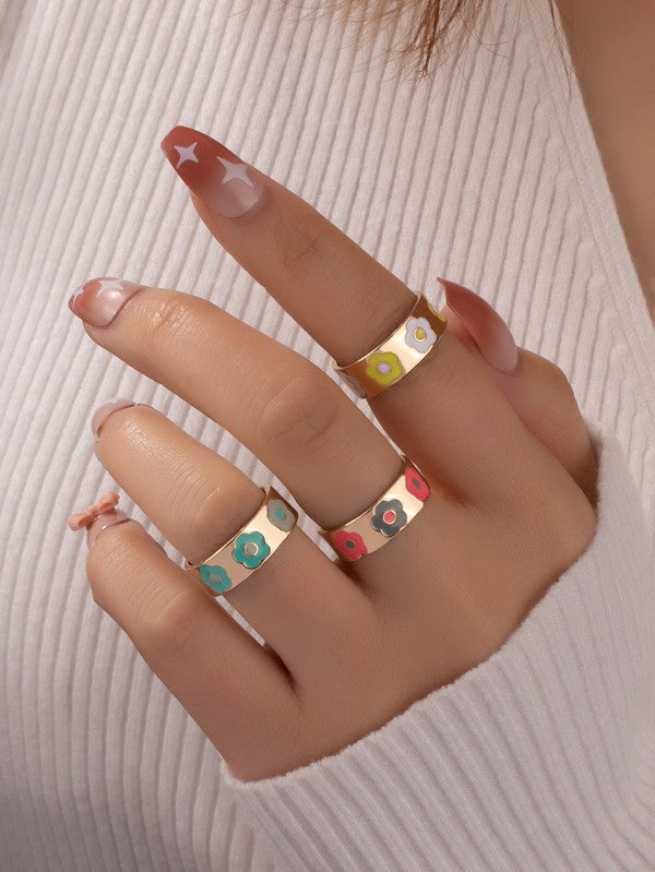 A hand with a white ribbed sleeve displays the "Set of three ring bands with flowers," showcasing vibrant gold rings adorned with colorful enamel flower designs on each finger. The long nails, painted in a pink and white pattern, beautifully complement these lively ring bands.
