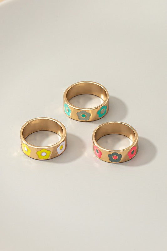 A hand with a white ribbed sleeve displays the "Set of three ring bands with flowers," showcasing vibrant gold rings adorned with colorful enamel flower designs on each finger. The long nails, painted in a pink and white pattern, beautifully complement these lively ring bands.