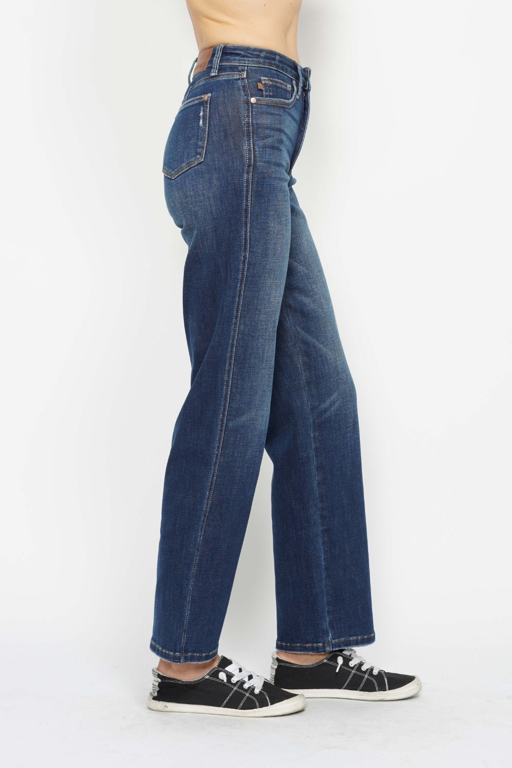 Person wearing Judy Blue Full Size High Waist Tummy Control Jeans in dark blue, paired with black and white sneakers, standing against a plain white background.