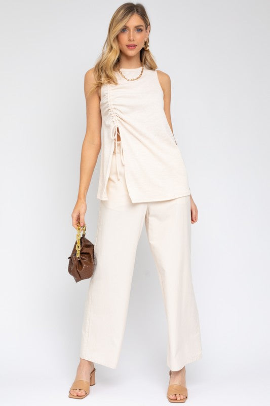 Dressed in a Sleeveless Ruching Top in beige and white pants, this woman carries a brown handbag, demonstrating a versatile style against a simple backdrop.