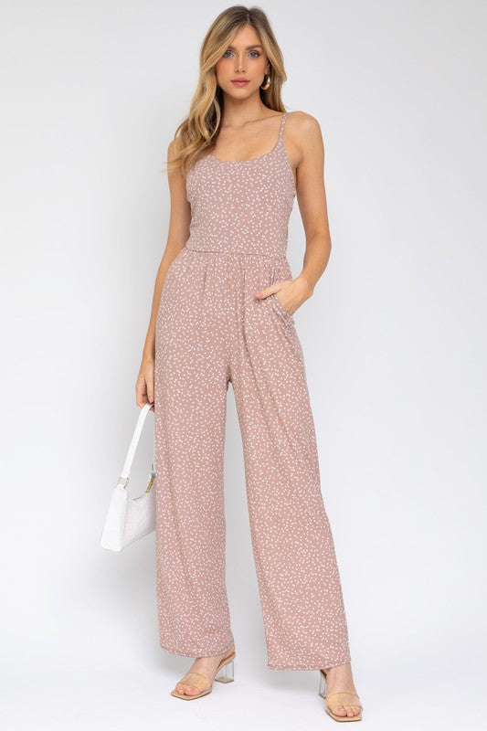 A woman models the Sleeveless Scoop Neck Wide Leg Jumpsuit, featuring a light pink pattern, against a plain white background while gently holding a strand of her hair with one hand.