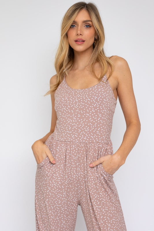 A woman models the Sleeveless Scoop Neck Wide Leg Jumpsuit, featuring a light pink pattern, against a plain white background while gently holding a strand of her hair with one hand.