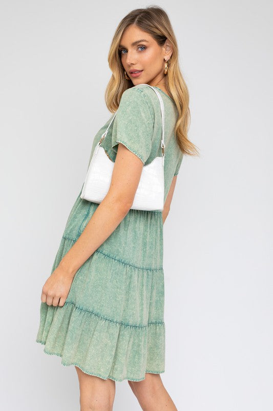 A woman in a Washed Baby Doll Mini Dress, featuring a vintage-inspired green design with a tiered skirt, holds a white handbag, perfectly poised for casual outings against a plain background.