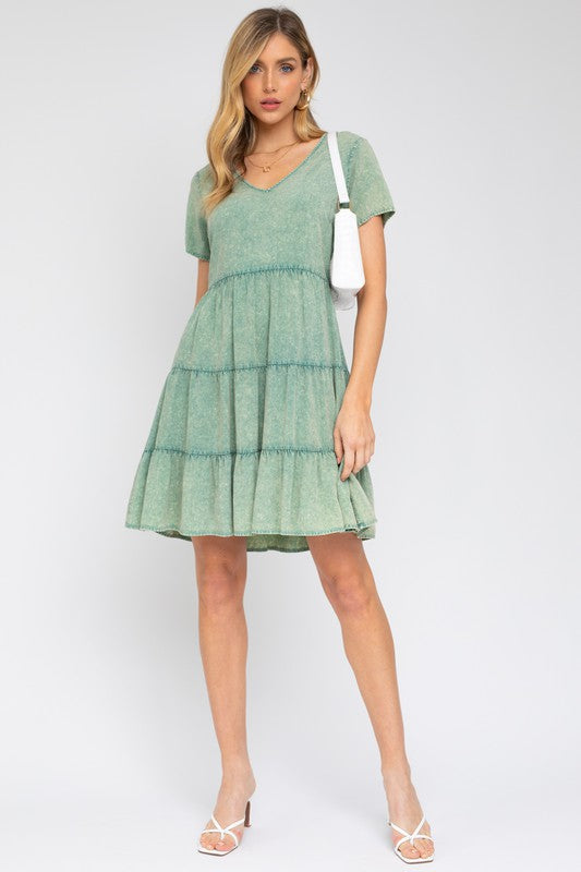 A woman in a Washed Baby Doll Mini Dress, featuring a vintage-inspired green design with a tiered skirt, holds a white handbag, perfectly poised for casual outings against a plain background.